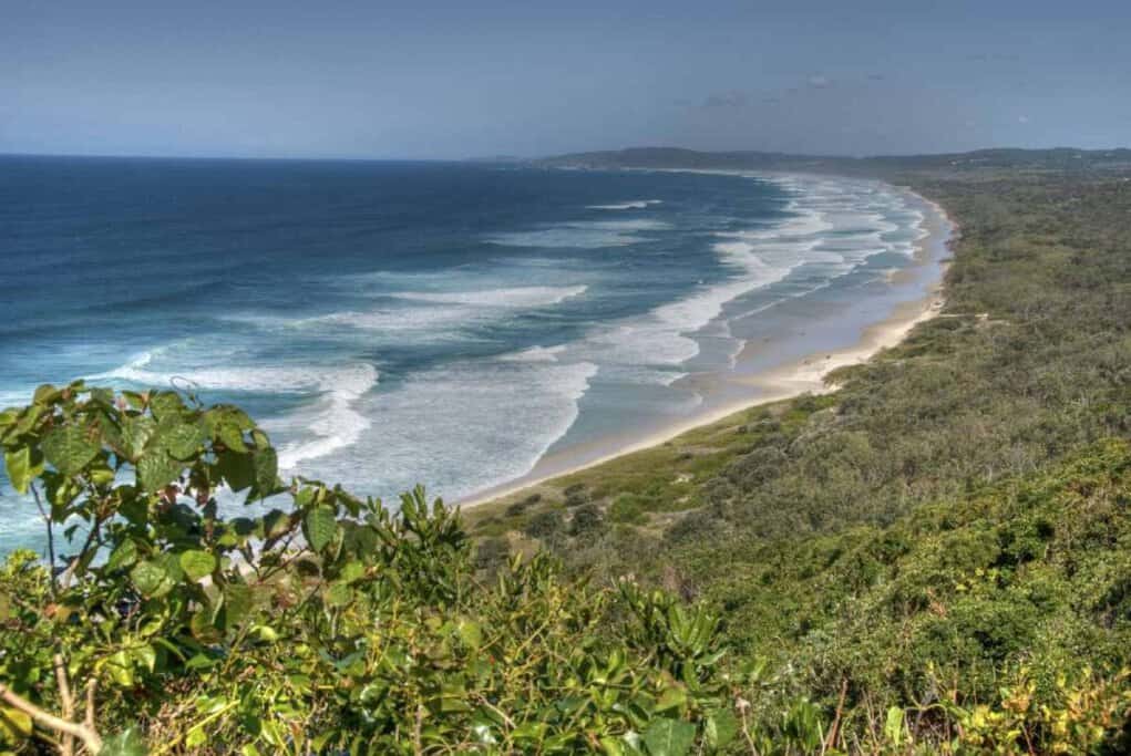 Panoramic view of a pristine, long sandy Byron Bay beach with gentle surf rolling onto the shore, framed by lush greenery, highlighting one of the best Australian surf spots.