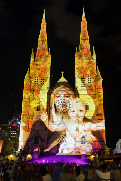 Crowds of people on the forecourt of St Mary's Cathedral Sydney CBD watch and enjoy the Christmas lights display. Madonna and Child one of the many visions on display on the church building sandstone facade at Christmas time.