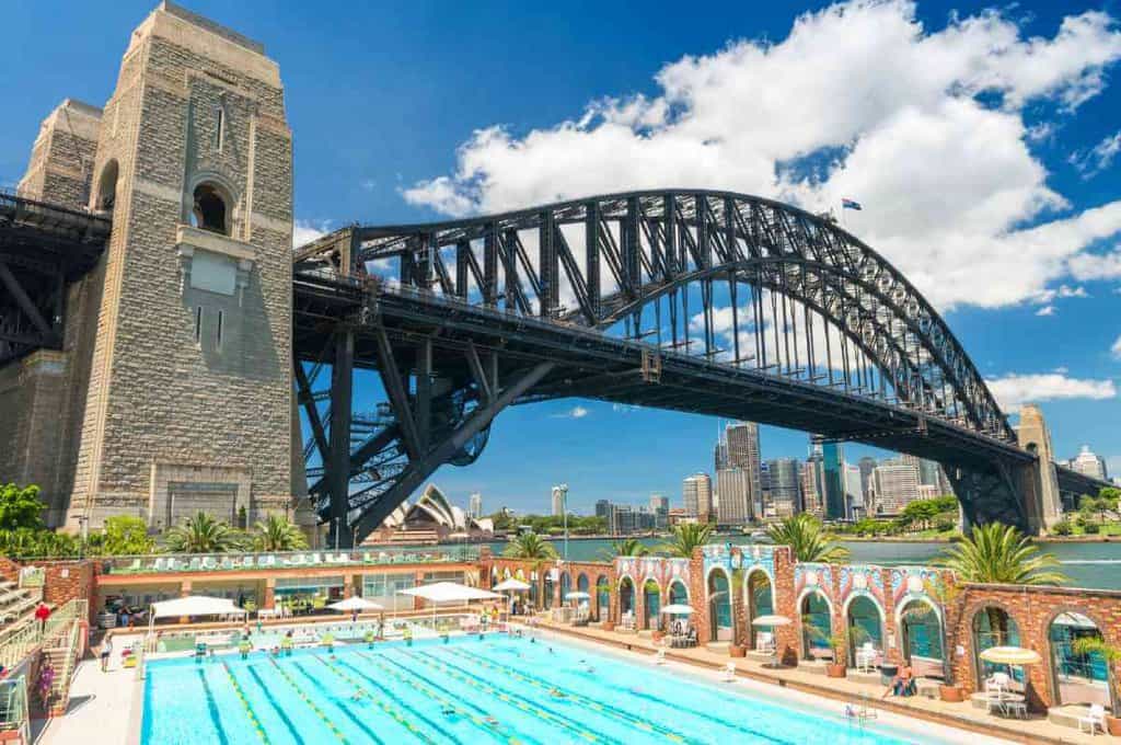 "Sydney Harbour Bridge towering over the North Sydney Olympic Pool, with its grand arch and steel cables, indicative of the significant weight the bridge can hold, set against a backdrop of the Sydney skyline and clear blue skies.