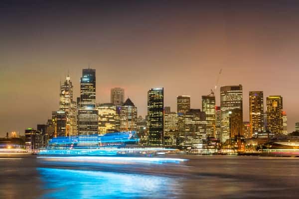 Panoramic night view of Sydney Harbour, with illuminated skyscrapers and a streak of ferry lights reflecting on the water, raising the question of time zones as explored in the context of Melbourne and Sydney's time alignment