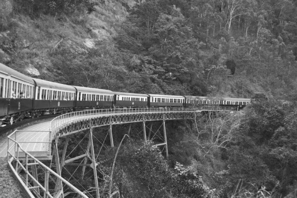 Historic Kuranda Scenic Railway train winding through lush rainforest and rugged terrain on its journey to Cairns. This iconic railway journey is a highlight in the comparison of attractions in Cairns or Airlie Beach.