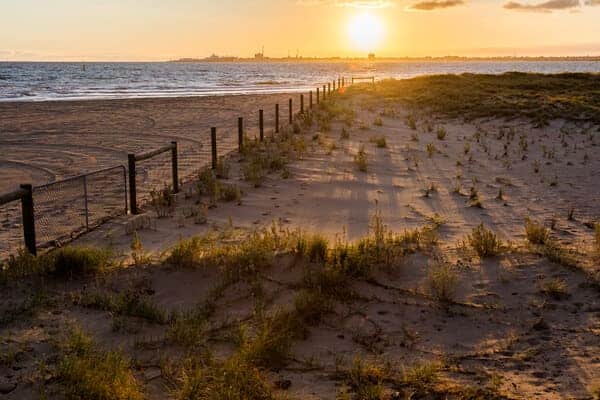 A serene Melbourne beach at sunset, with the sun casting long shadows over the sandy path and dunes, while a calm sea stretches into the distance. This tranquil scene contrasts with the theme of the blog post, illustrating the calm before the often unpredictable weather shifts in Melbourne.