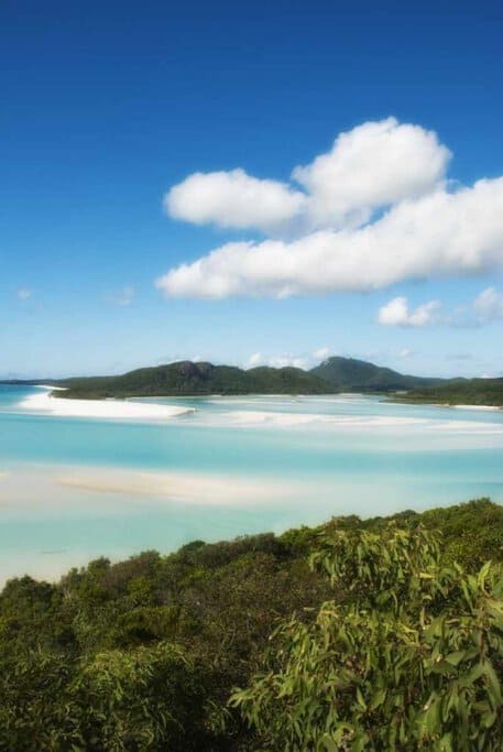 A serene view of Whitehaven Beach in the Whitsundays, showcasing the crystal-clear turquoise waters contrasted with pristine white sands, flanked by lush greenery, under a bright blue sky with fluffy clouds—highlighting the natural beauty that distinguishes the Whitsundays in the 'Port Douglas vs. Whitsundays' debate.