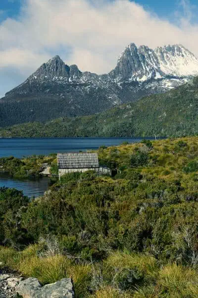 The iconic Cradle Mountain in Tasmania looms in the background with a dusting of snow on its peaks, overlooking the historic boatshed at Dove Lake. This striking image highlights the captivating destinations explored during Cradle Mountain walking tours.