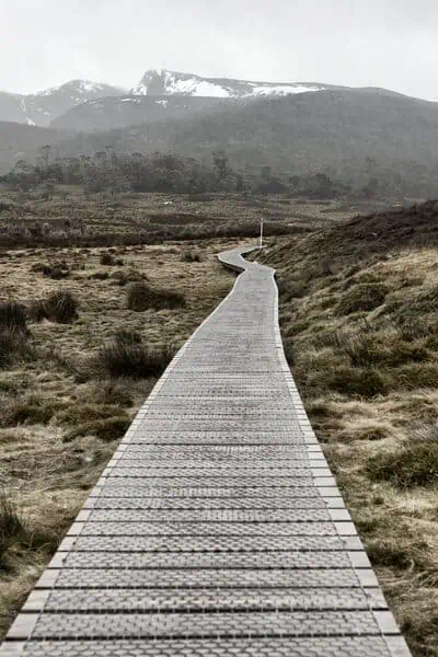 A winding boardwalk leads adventurers through the rugged terrain of Cradle Mountain, Tasmania, with the mountain's snowy peaks in the distance. This path is part of the diverse walking tracks that make up the Cradle Mountain hiking tours, offering an accessible route through the wild landscape.
