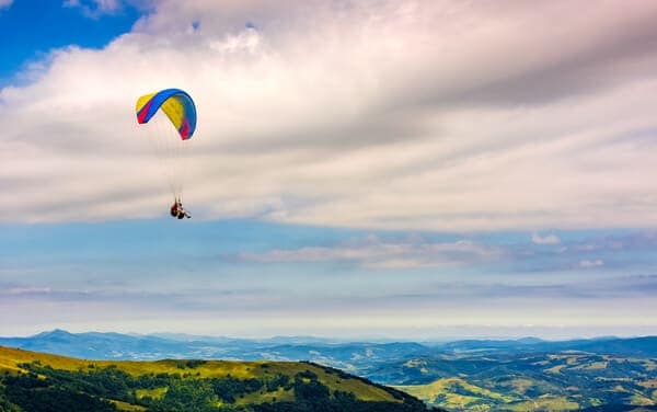 A paraglider soars high above rolling hills under a dramatic sky, offering an experience as freeing as bungee jumping on Sydney Harbour Bridge.