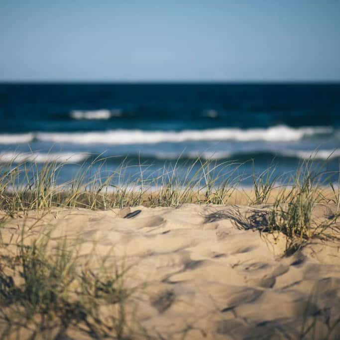 Close-up of sand dunes with sparse grass overlooking a blurred background of ocean waves, ideal for surfing in Australia.