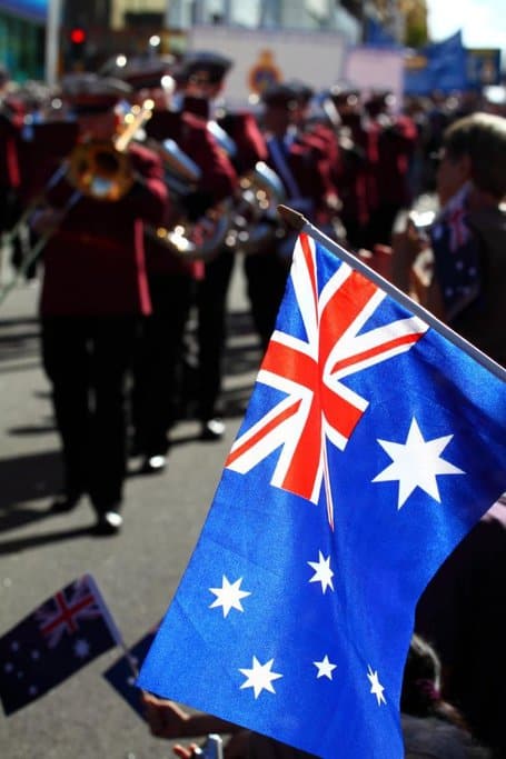 An Australian flag waving in the foreground, with a parade of marching band members in the background. This scene captures the festive atmosphere of events like ANZAC Day, where quirky Aussie traditions, including parades and flag displays, take centre stage.