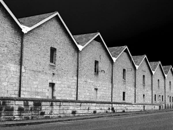 Black and white image of the historical buildings at The Rocks in Sydney, lined up with their pointed gable roofs against the night sky. The mysterious ambiance of the area is a key feature of the spooky narratives explored in Sydney ghost tours.
