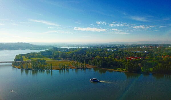 A panoramic view of Canberra from above, showing Lake Burley Griffin and its surrounding parklands with a boat cruising on the water, illustrating the tranquil beauty encountered on Canberra tours from Sydney.