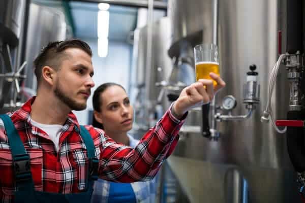 Two brewers critically assessing the quality of a golden beer sample in a glass, amidst the stainless steel tanks of a brewery in Brisbane, showcasing the expertise involved in the brewery tours.