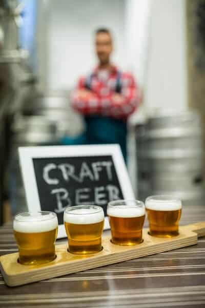 A selection of four craft beers presented on a wooden tray in the foreground, with a brewer standing proudly behind, near a 'Craft Beer' sign, highlighting the diversity of offerings on brewery tours in Brisbane.