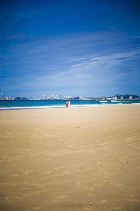 Two people strolling along a vast, sandy beach with the Gold Coast skyline in the background under a clear blue sky, showcasing popular surfing spots in Australia.