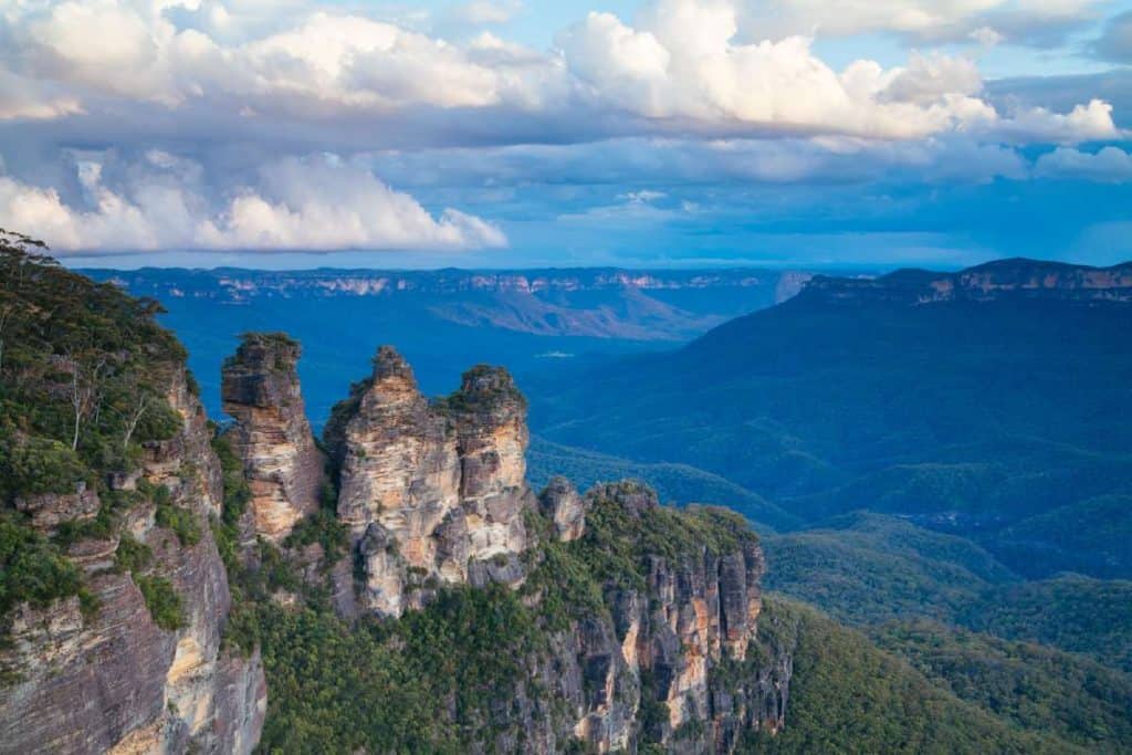 View of the Three Sisters rock formation surrounded by the expansive blue-hued valleys of the Blue Mountains, New South Wales, Australia. Showcases a famous natural landmark, connecting to a blog post about Australia's iconic destinations.