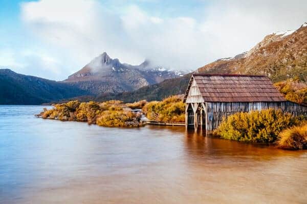The historic boatshed at Dove Lake, with Cradle Mountain enshrouded by clouds in the background, as seen on a serene Tasmanian day. This picturesque scene is a highlight for many exploring the natural wonders on Cradle Mountain walking trails.