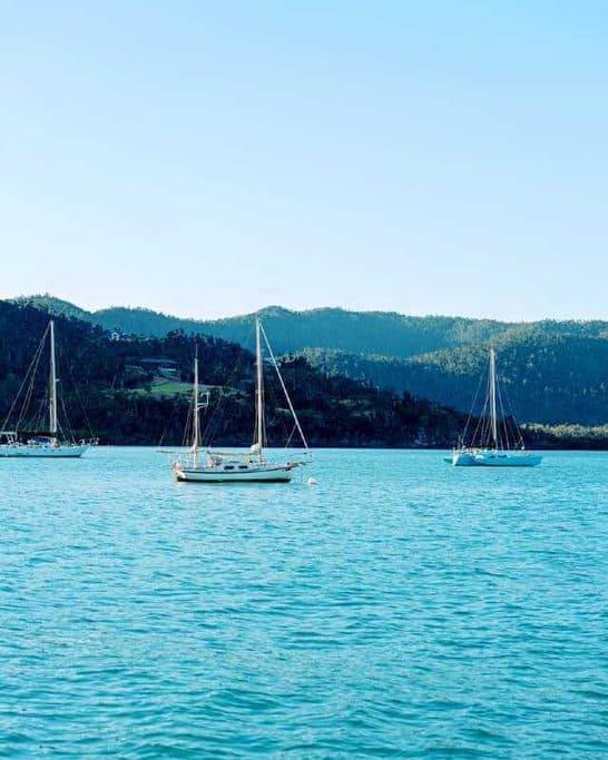 Sailboats anchored in the tranquil waters of Airlie Beach, with lush green hills in the background. This idyllic scene represents the serene sailing opportunities that are compared in the Cairns and Airlie Beach travel guide.