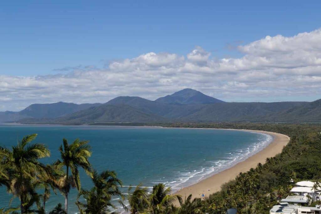 A sweeping view from Trinity Bay Lookout in Port Douglas, Queensland, showcasing a sweeping curve of golden sandy beach lined with palm trees, with the Coral Sea's azure waters on one side and a backdrop of majestic green mountains under a sky dotted with clouds. This coastal panorama underscores the vibrant allure of Port Douglas in the 'Port Douglas vs Whitsundays' exploration.