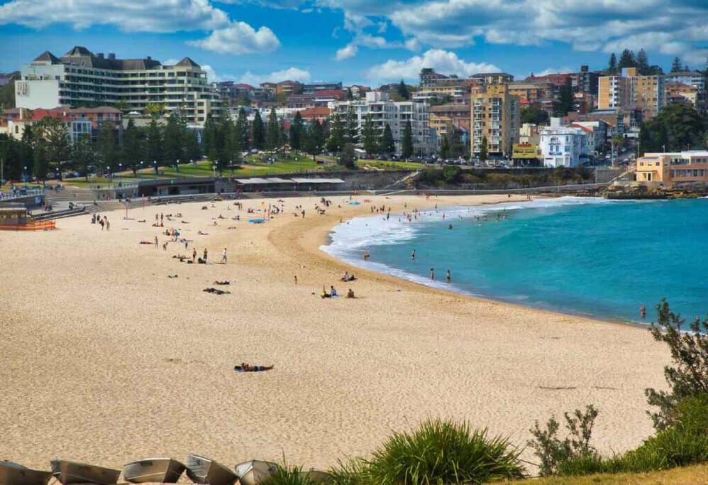 Busy day at Manly Beach with beachgoers and swimmers enjoying the calm blue waters, surrounded by the urban skyline, making it one of the best places to surf in Australia.