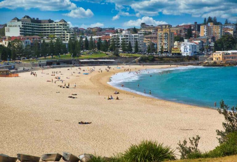 Busy day at Manly Beach with beachgoers and swimmers enjoying the calm blue waters, surrounded by the urban skyline, making it one of the best places to surf in Australia.
