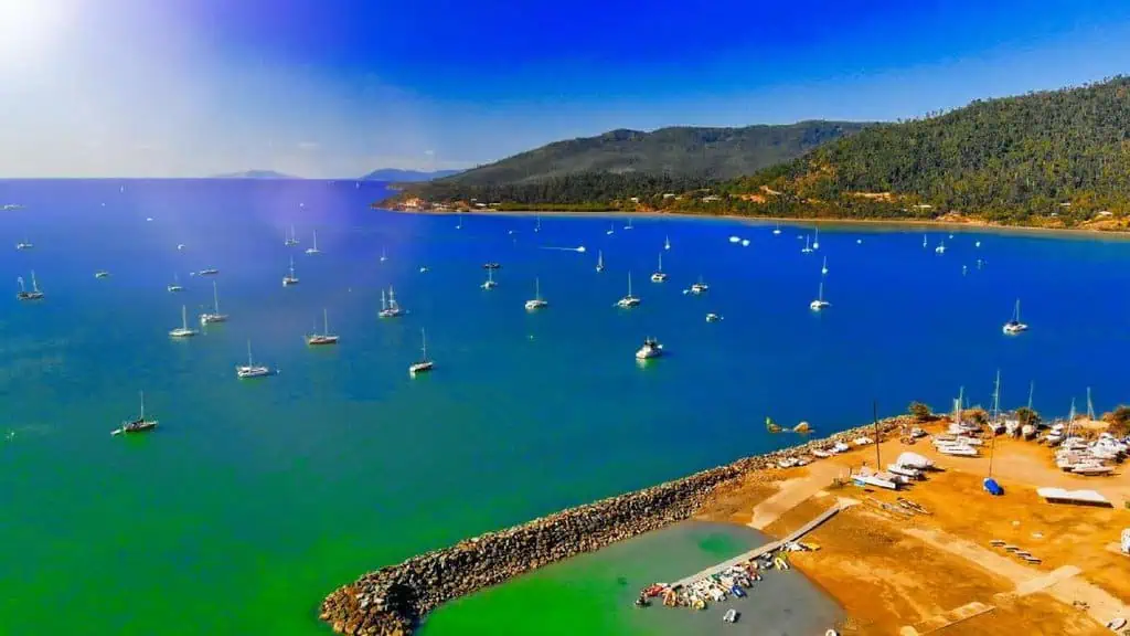 Aerial view of numerous sailboats anchored in the clear blue waters of Airlie Beach, with lush hills in the background. This beautiful marine vista is a key attraction discussed in the Airlie Beach vs Cairns travel comparison.