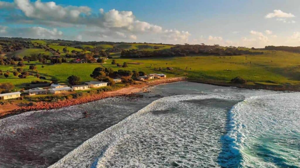 Aerial view of a tranquil bay with frothy waves ideal for surfing, set against a lush, green landscape and quaint beachfront properties, epitomizing Australia beach surfing.
