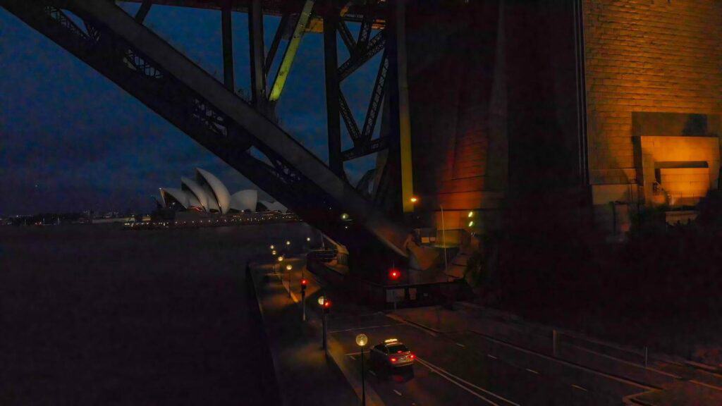 Aerial night view from under the Sydney Harbour Bridge with the Sydney Opera House in the distance, street lights casting a warm glow on the road. This hauntingly beautiful scene is reminiscent of the mysterious vibes found on Sydney ghost tours.