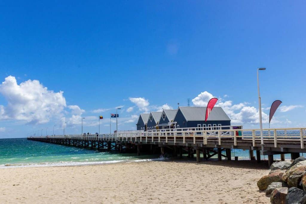 View of Busselton Jetty extending into the ocean under a bright blue sky with clouds, in Western Australia. This jetty is an example of one of the longest jetties in Australia, often visited by tourists for its picturesque views and historical significance.