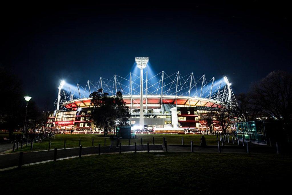 The Melbourne Cricket Ground (MCG) illuminated at night, home to thrilling AFL matches and a hub for quirky Australian sporting traditions. This iconic venue is where fans gather to enjoy the game, indulge in classic footy snacks like meat pies, and embrace the unique atmosphere of Aussie culture."