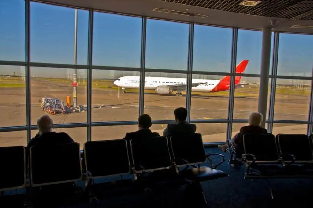 Passengers seated in an airport terminal, looking at a Qantas plane on the tarmac preparing for takeoff. This image relates to the question 'How Far Is Adelaide From Sydney?' by depicting a common mode of travel between the two cities, with flights being a convenient option for the journey