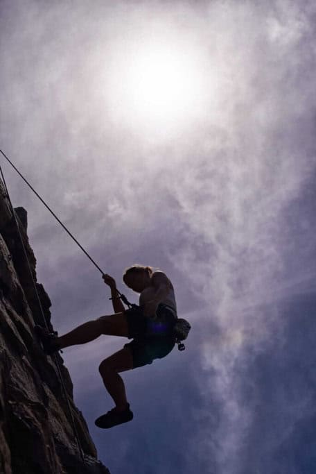 A silhouette of a rock climber abseiling down a cliff, set against a hazy sun, captures the adventurous spirit akin to bungee jumping at the Sydney Harbour Bridge.