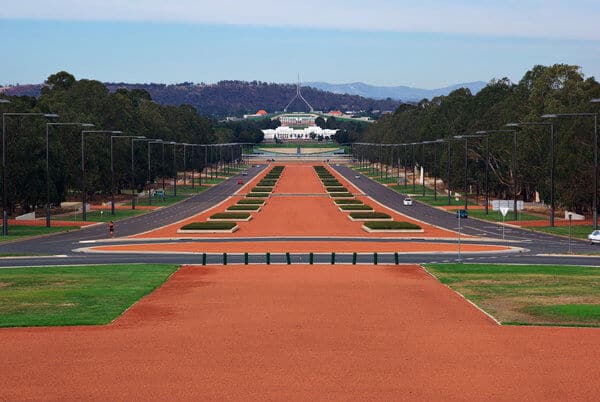 iew of the Parliamentary Triangle in Canberra, featuring the Parliament House at the end of a long, red-gravelled ceremonial avenue, surrounded by lush greenery. This image relates to the post by showcasing a popular stop on Canberra tours from Sydney.