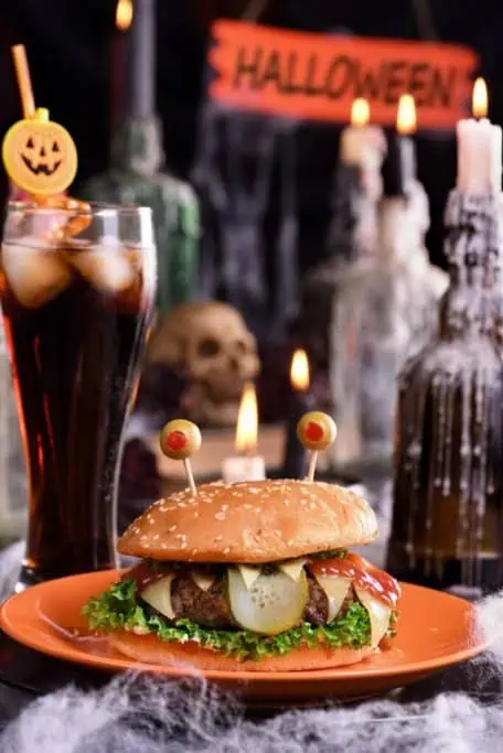 A Halloween-themed burger with olive 'eyes' and a spooky drink in the background, surrounded by candles and skull decor. This creative meal sets the mood for a fun Halloween party in Sydney, where themed food and decorations play a big part in the festive celebrations.