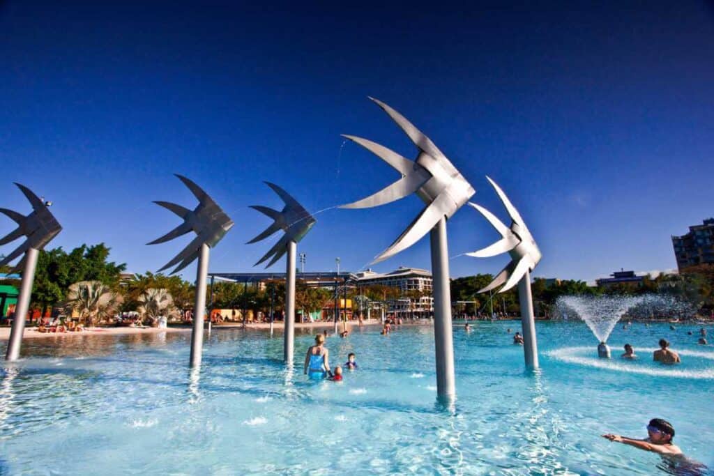 Families enjoying the water features beneath large metallic fish sculptures at Cairns Esplanade, a popular public space comparing the relaxed urban atmosphere of Cairns with the natural splendour of the Whitsundays