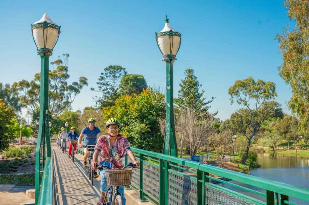 A group of people enjoying an Adelaide e-bike tour, crossing a scenic bridge with lush greenery and the River Torrens in the background. This image highlights the relaxing and picturesque experience of exploring Adelaide's sights on an electric bike.