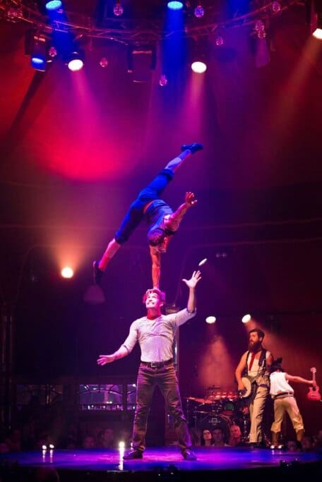 Two male acrobats performing an impressive balancing act under colourful stage lights at the Adelaide Fringe Festival, with live musicians in the background — showcasing the thrilling entertainment found at Epic Australian Festivals.