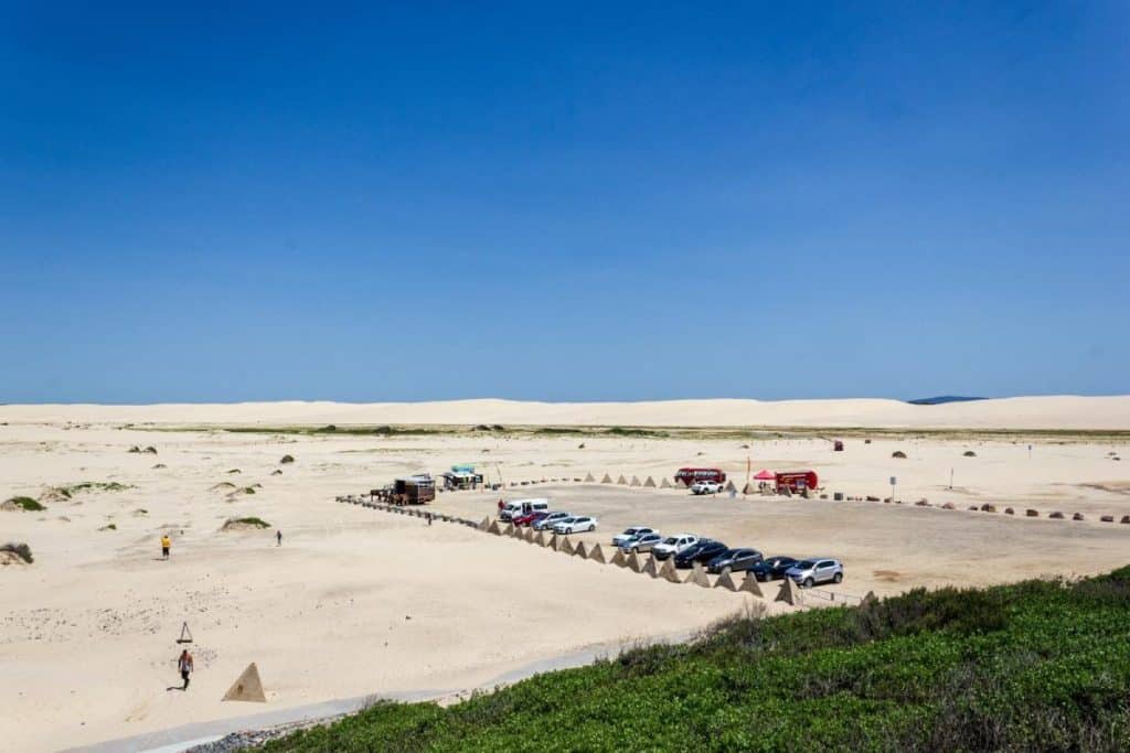 A wide view of Stockton Sand Dunes at Anna Bay with parked cars, food trucks, and tourists scattered across the golden sand under a clear blue sky – a popular stop for sandboarding and off-road tours during a road trip from Sydney to Byron Bay.