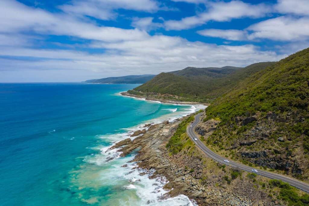 Aerial view of the winding Great Ocean Road hugging the rugged coastline with turquoise waters and lush hills under a blue sky — a scenic route often explored when planning how to get to Melbourne from Adelaide on a coastal road trip.