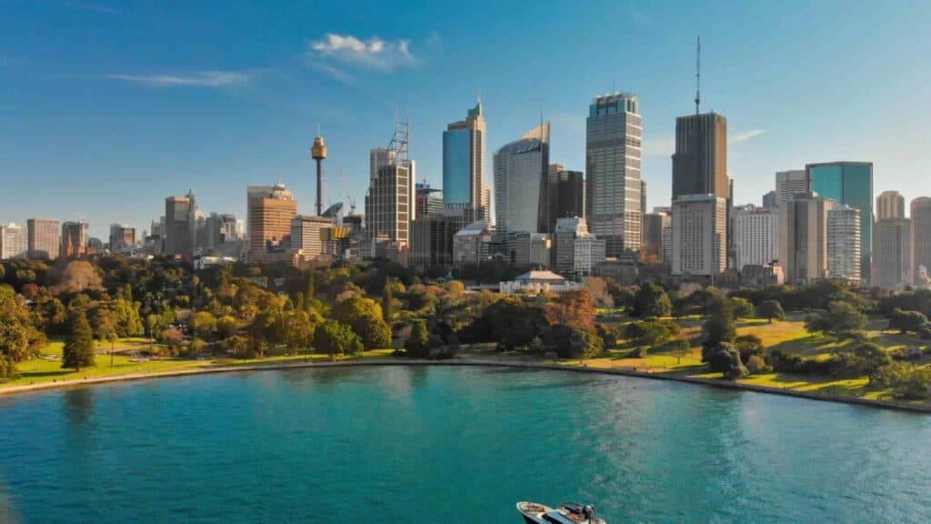Aerial view of Sydney Harbour, showcasing the vibrant city skyline with iconic buildings and lush greenery along the waterfront under a clear blue sky. This image captures the dynamic energy of Sydney during summer, highlighting it as a premier urban destination in Australia.