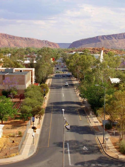 View of a central street in Alice Springs, lined with greenery and framed by the rugged West MacDonnell Ranges in the distance, showcasing the unique blend of urban and natural landscapes experienced when travelling to Alice Springs.