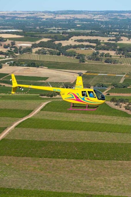 A yellow Robinson R44 helicopter flying over the patchwork of vineyards and fields in the Barossa Valley, showcasing the picturesque landscape. This image exemplifies the unique experiences offered by helicopter hire Adelaide, allowing visitors to witness the expansive beauty of South Australia from an exceptional aerial perspective.