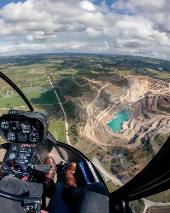 View from inside a helicopter cockpit, showcasing the pilot's perspective as they soar over a scenic landscape featuring lush greenery and a striking quarry with turquoise water. This vivid scene from helicopter tours Adelaide highlights the dramatic and diverse terrain accessible on these aerial adventures.