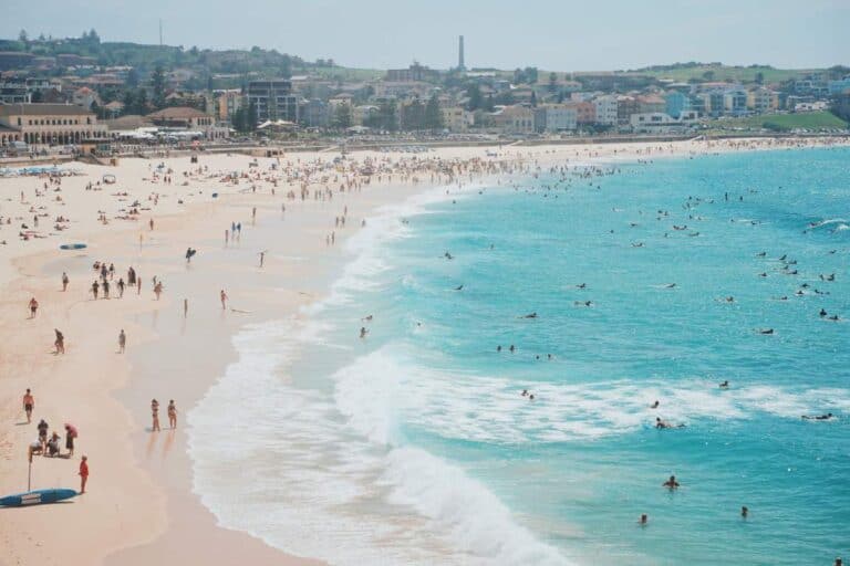 Busy day at Bondi Beach with swimmers and sunbathers enjoying the sun and surf, set against the backdrop of the nearby town. This vibrant scene captures the essence of a classic summer day in Australia, showcasing Bondi Beach as a must-visit destination for beach lovers.