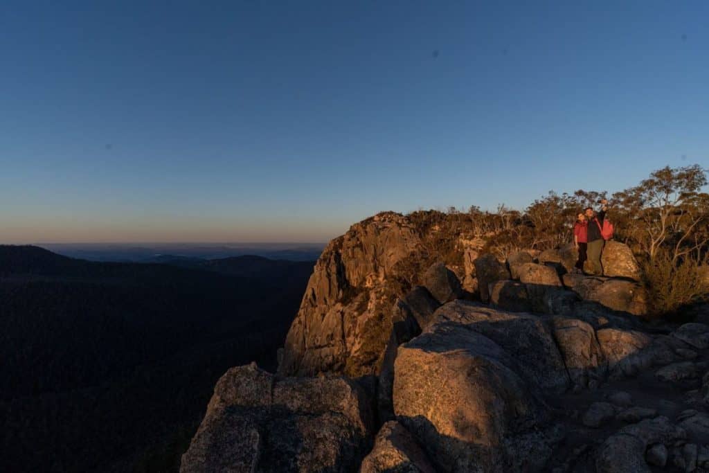 Two hikers standing atop Booroomba Rocks at sunset, taking in panoramic views of the Namadgi National Park. This rugged lookout is a hidden hiking gem in the ACT, offering breathtaking vistas and a rewarding climb for adventurers seeking an off-the-beaten-path experience near Canberra.