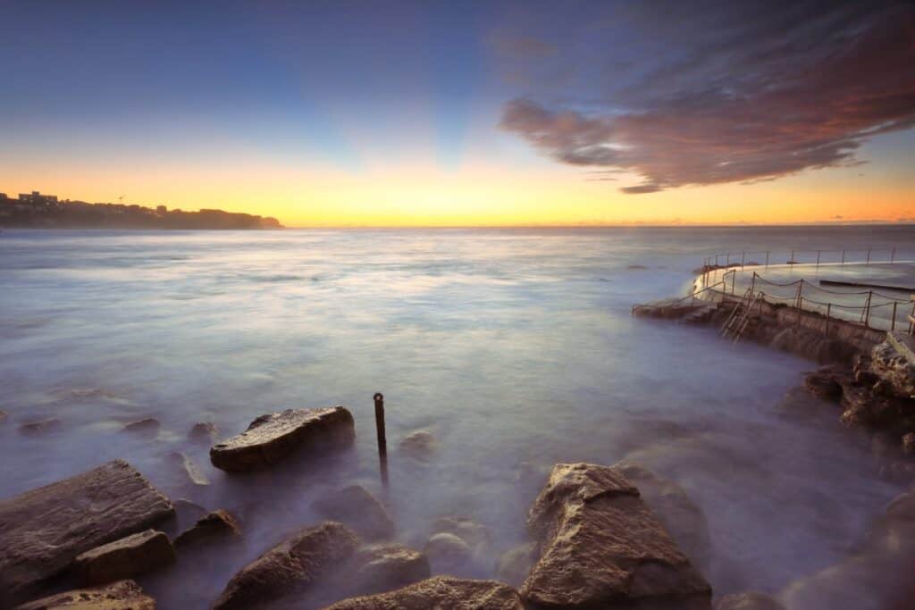 Bronte Beach at sunrise, showing a serene view with smooth waves and rocky formations along the shoreline. Despite its scenic beauty, Bronte Beach is sometimes considered one of the worst beaches in Australia for its hazardous rip currents and overcrowded conditions during peak times.