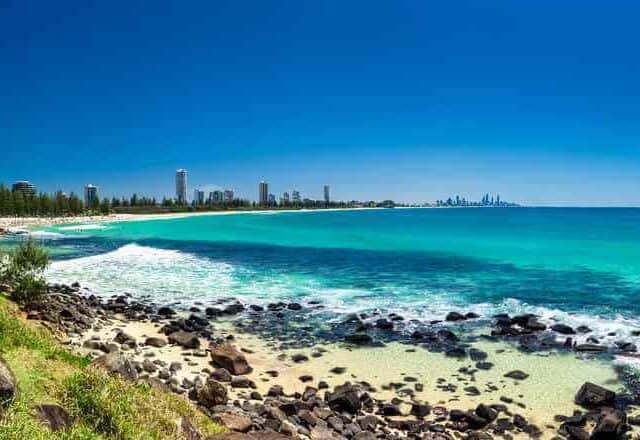 A panoramic view of Burleigh Heads beach with turquoise waters and a rocky shoreline in the foreground, framed by lush greenery and with Gold Coast high-rises in the distance, showcasing the diverse beauty of beaches in Australia.
