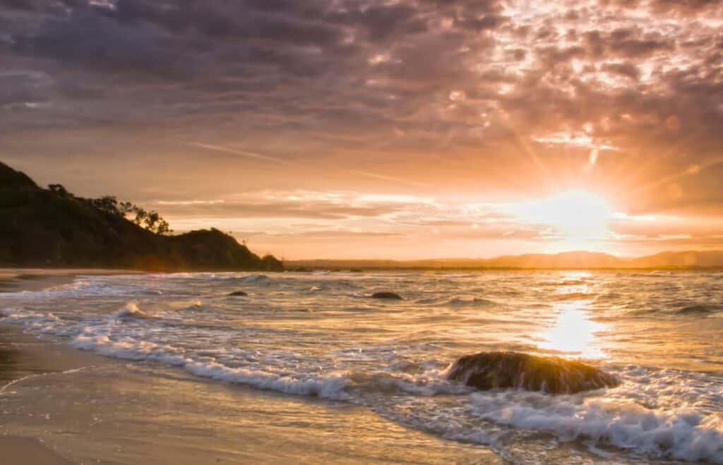 Golden sunset over the beach at Byron Bay, with soft waves reflecting the sun’s glow as it sets behind the hills. This image highlights one of the best locations for iconic sunset views in Australia, supporting the blog post about must-see sunset spots.