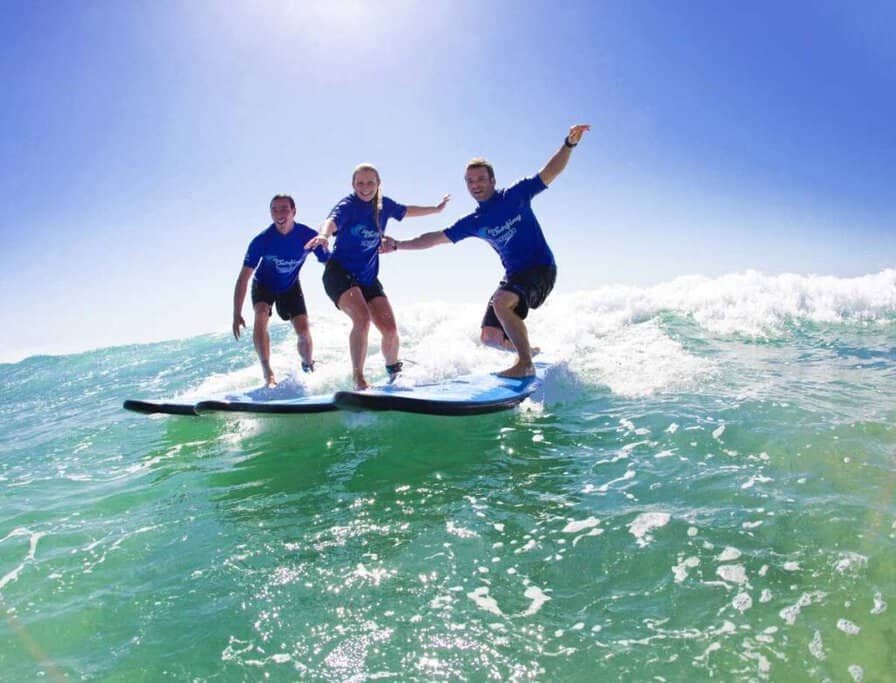 Three people taking a surf lesson ride a wave together under a clear blue sky, embodying the fun and excitement of learning to surf when you travel to Byron Bay.