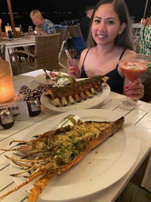 The author, Kate, at a beachfront restaurant in the Gili Islands, enjoying local seafood cuisine, with a grilled lobster and skewered meat on the table, and a cocktail in hand. This dining experience highlights the culinary delights as one of the many things to do in Gili Islands.