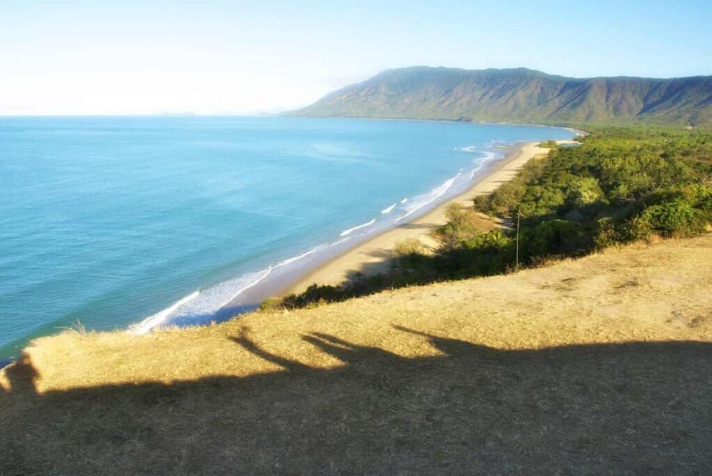 Expansive view of a sunlit beach along the Cairns coast, with calm blue waters stretching towards distant mountains. This image highlights the serene beauty of summer in Australia, showcasing Cairns as an ideal destination for a coastal retreat.