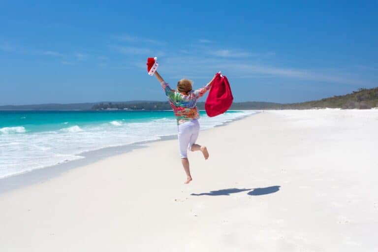 A person running along a beach holding a Santa hat and red bag, celebrating Christmas under the summer sun. One of the fun Australian Christmas facts is that many people celebrate the holiday outdoors, making the most of the warm weather and stunning beaches.